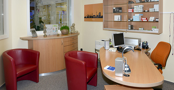 Modernes Beratungszimmer mit Schreibtisch, Computer, Regalen und roten Sesseln.